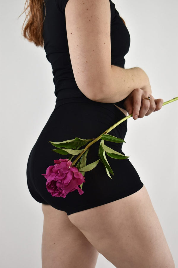 A woman wearing the Her Bloom Shortie in black, holding a pink peony against her hip