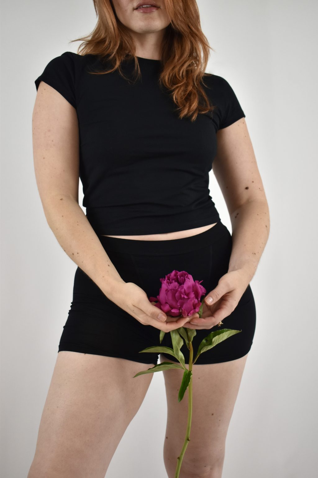 A woman in Her Bloom period underwear holding a magenta peony cupped in both hands at her lower stomach