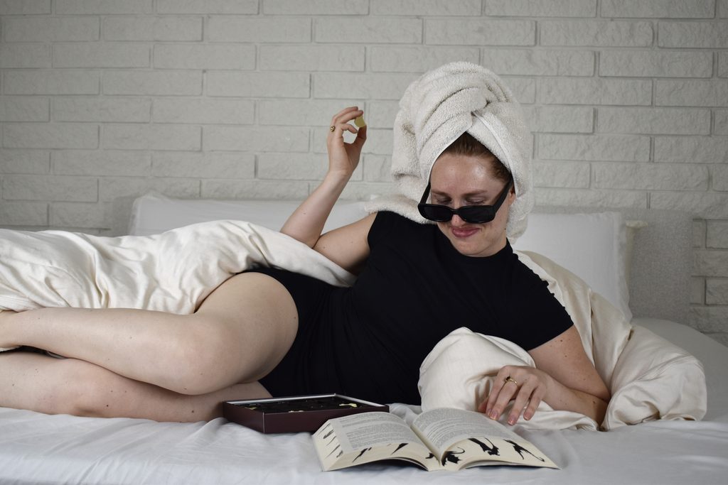 A woman relaxing on a bed in Her Bloom period underwear, with a book and a box of chocolates