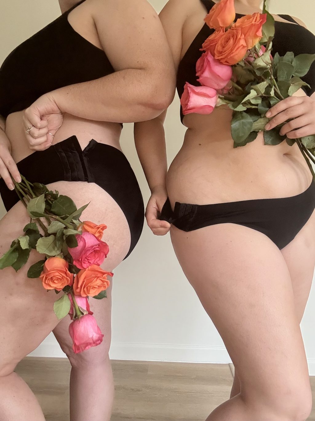 Two women standing side by side in black Her Bloom period underwear, holding pink and orange roses, with the adjustable side closures visible