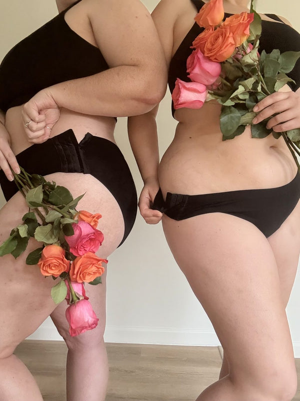 Two women standing side by side in black Her Bloom period underwear, holding pink and orange roses, with the adjustable side closures visible