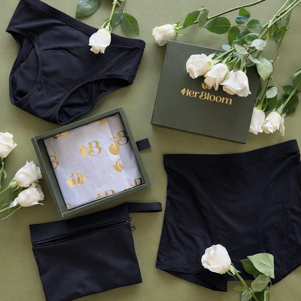 A flat lay of Her Bloom products in black on a sage background, including the Shortie, the Keep it Brief, Brief, a travel bag, and an open gift box with gold printed tissue paper, surrounded by white roses