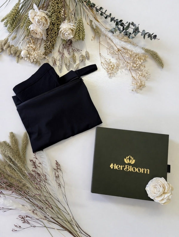 The Her Bloom Travel Bag in black beside a sage gift box with the gold Her Bloom logo, surrounded by dried flowers