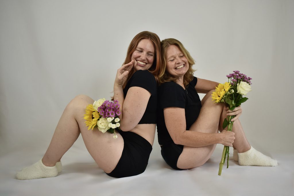 Two women sitting back to back and laughing together in Her Bloom period underwear, holding bunches of flowers