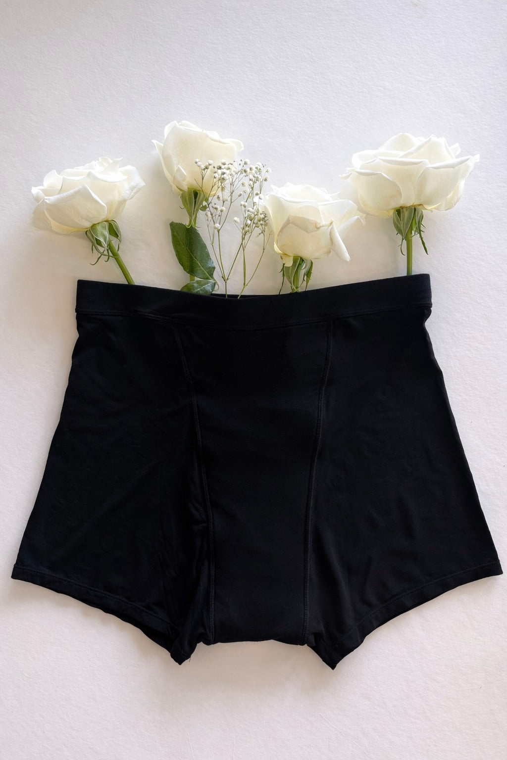 A pair of Her Bloom Shortie period underwear in black, laid flat with white roses on a soft neutral background