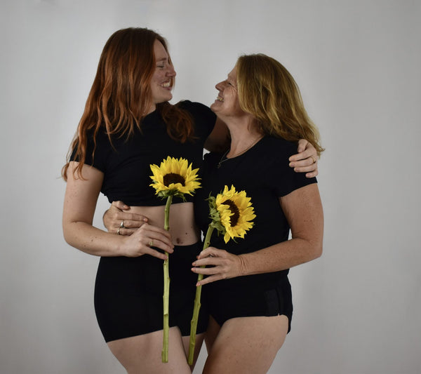 Two women of different ages laughing together, wearing black Her Bloom period underwear and black tees, each holding a sunflower