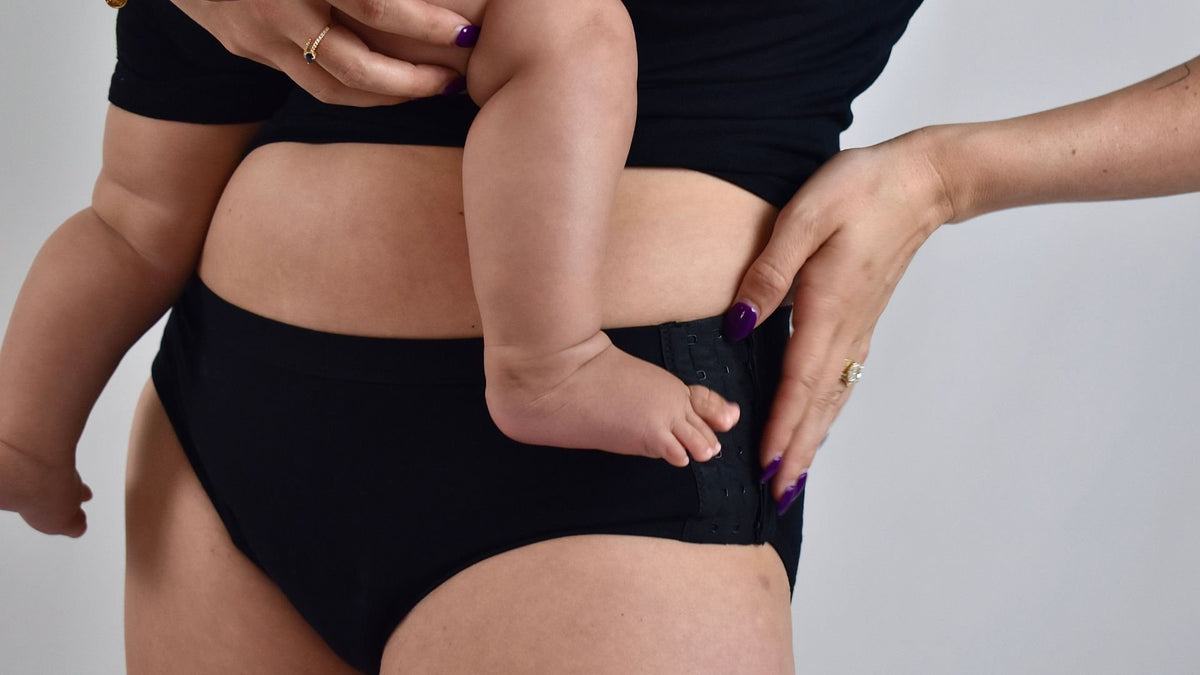 A woman in black Her Bloom period underwear holding a baby on her hip