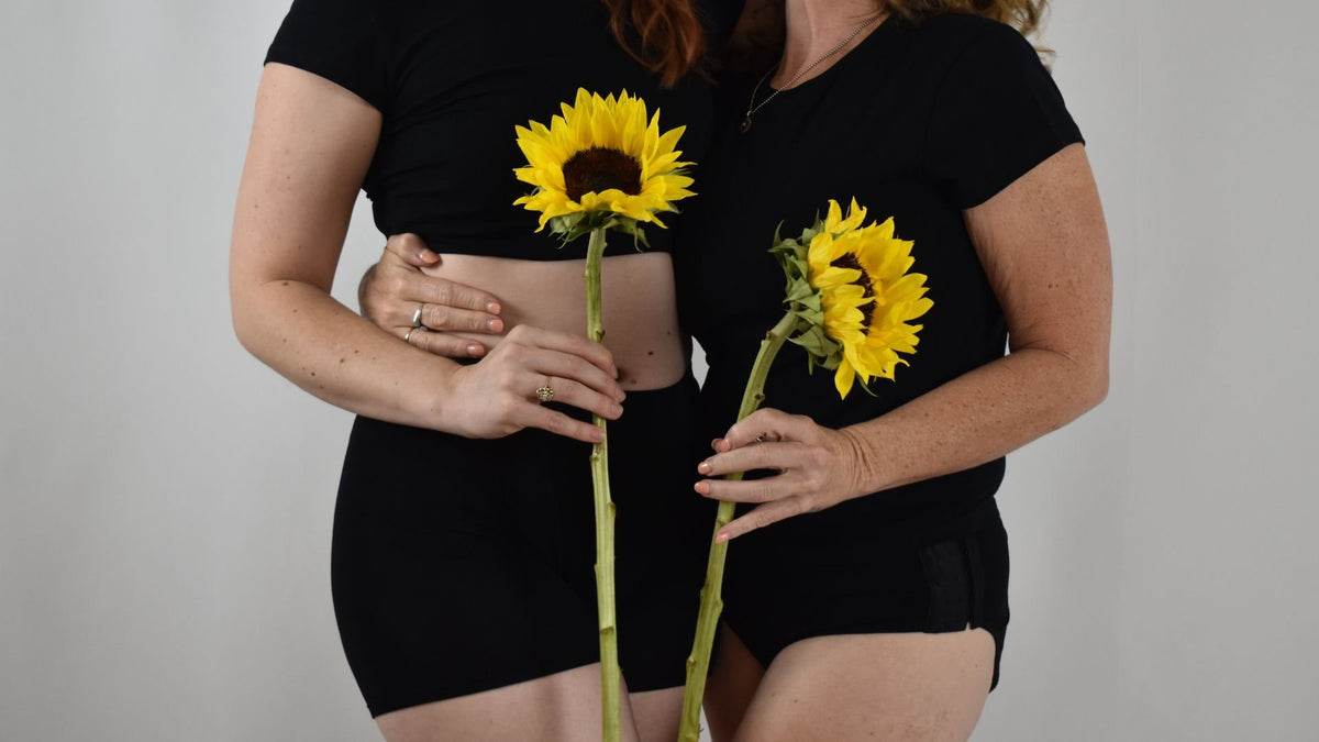 Two women in black Her Bloom period underwear, each holding a sunflower