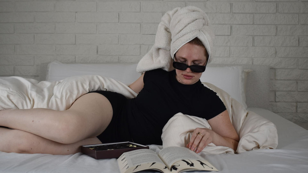 A woman in Her Bloom period underwear lounging in bed with a book, hair wrapped in a towel