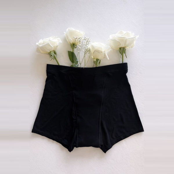 A pair of Her Bloom Shortie period underwear in black, laid flat with white roses on a soft neutral background
