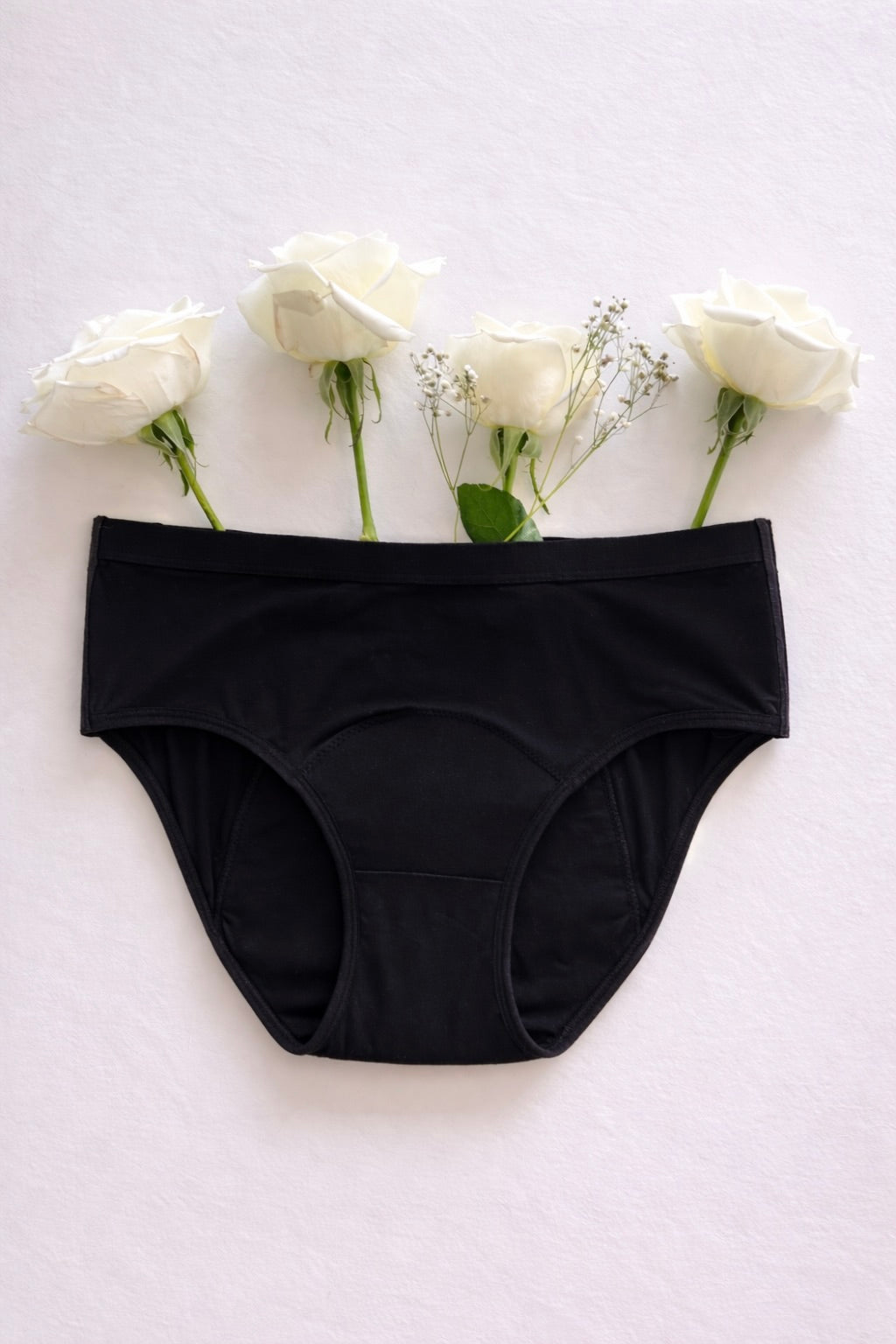 A pair of Her Bloom Keep it Brief period underwear in black, laid flat with white roses on a soft neutral background